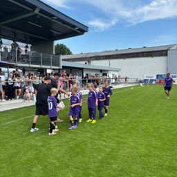 Eine Gruppe von Kindern in lila Fußballtrikots wird auf einem Feld von Erwachsenen begrüßt. Zuschauer schauen von einer Tribüne und Tribüne zu.