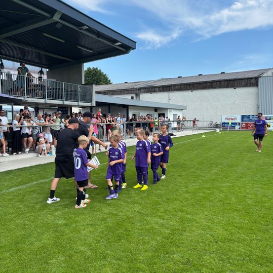 Eine Gruppe von Kindern in lila Fußballtrikots wird auf einem Feld von Erwachsenen begrüßt. Zuschauer schauen von einer Tribüne und Tribüne zu.