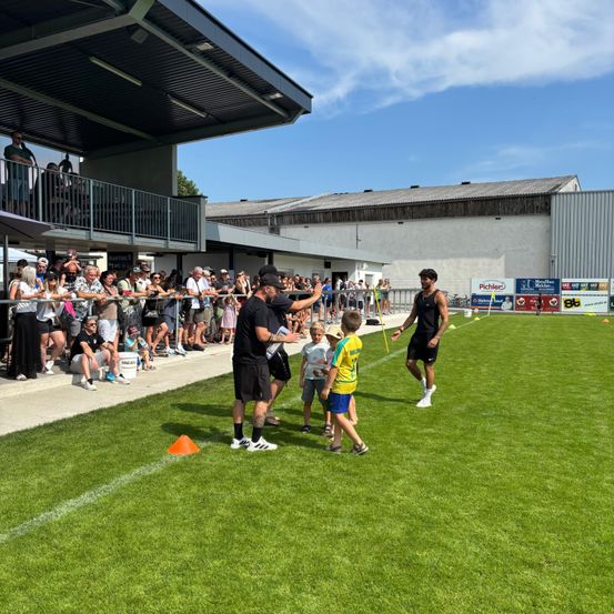 Ein Fußballspiel im Gange mit Spielern auf dem Feld, ein Trainer interagiert mit Kindern und Zuschauer schauen von den Tribünen aus bei einem sonnigen Tag zu.