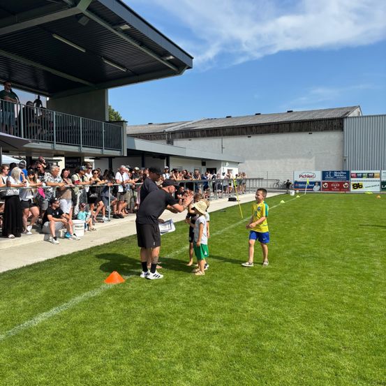 Ein Fußballtrainer gibt jungen Spielern auf dem Feld Anweisungen, während Zuschauer von den Tribünen zusehen. Das Feld ist mit Kegeln und weißen Linien markiert.