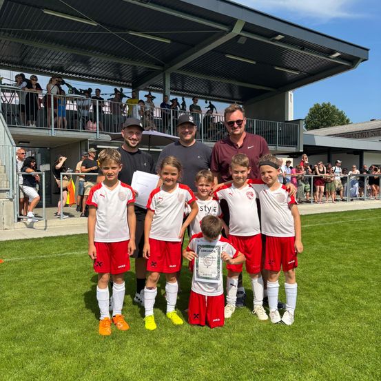 Eine Gruppe junger Fußballspieler, die weiße und rote Trikots tragen, stehen auf dem Feld mit Erwachsenen. Sie halten Urkunden in der Hand und posieren für ein Foto. Dahinter beobachtet eine Menge Menschen an den Seitenlinien.