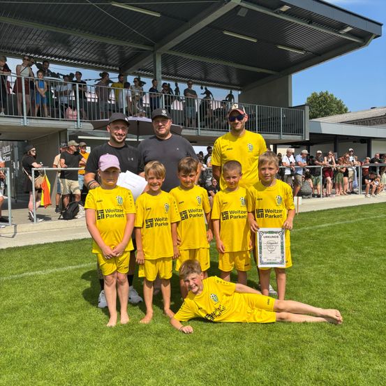 Eine Gruppe von Kindern in gelben Uniformen, wahrscheinlich eine Fußballmannschaft, posiert mit ihrem Trainer für ein Foto. Sie halten ein Zertifikat und sind auf einem Feld. Zuschauer beobachten von hinter einer Mauer.