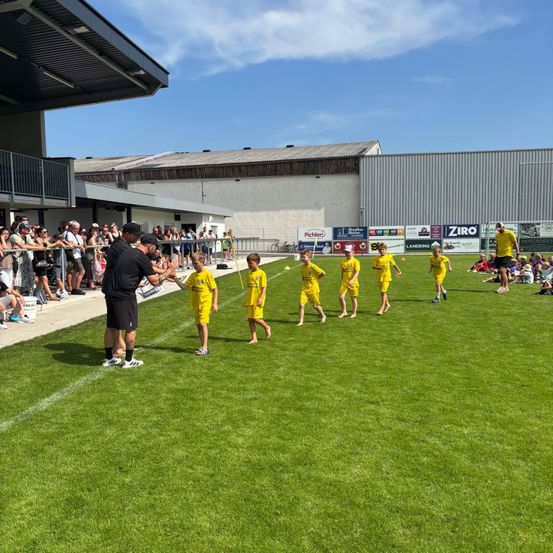 Eine Gruppe von Kindern in gelben Uniformen läuft auf einem Feld, während ein Mann in Schwarz Anweisungen gibt. Zuschauer beobachten von den Tribünen aus.