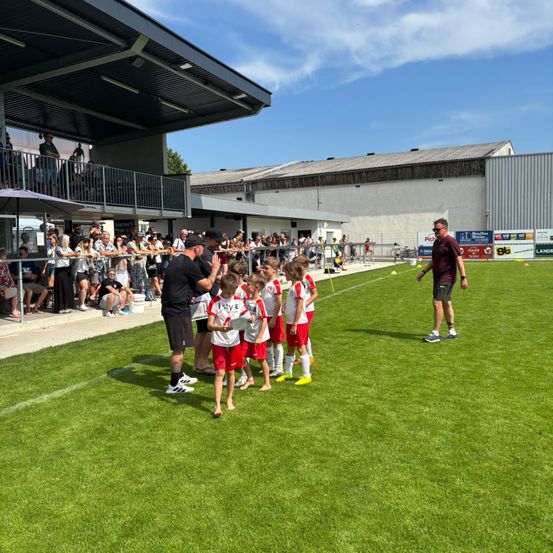 Ein Fußballspiel mit Spielern auf dem Feld, Trainern und Zuschauern. Die Spieler tragen rote Uniformen, und der Platz ist gut gepflegt.