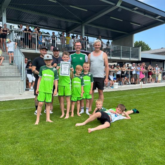 Eine Fußballmannschaft in grünen Trikots steht auf einem Rasen. Erwachsene und Kinder halten Urkunden und lächeln für die Kamera, mit einer Menge im Hintergrund.