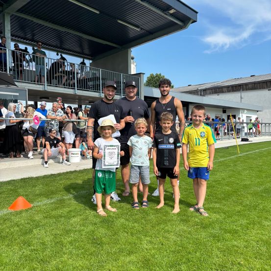 Eine Gruppe von Menschen steht auf einem Feld, wobei vier Kinder ein Zertifikat halten. Dahinter stehen zwei Erwachsene, einer trägt einen Hut. Zuschauer beobachten von einem eingezäunten Bereich aus. Bäume und Gebäude sind im Hintergrund.