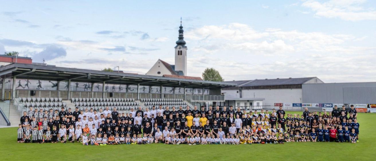 Eine große Gruppe von Fußballspielern, Trainern und Kindern posiert für ein Foto auf einem Feld, mit einer Kirche und einem Stadion im Hintergrund.