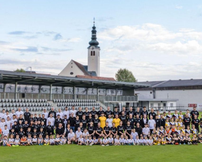 Eine große Gruppe von Fußballspielern, Trainern und Kindern posiert für ein Foto auf einem Feld, mit einer Kirche und einem Stadion im Hintergrund.