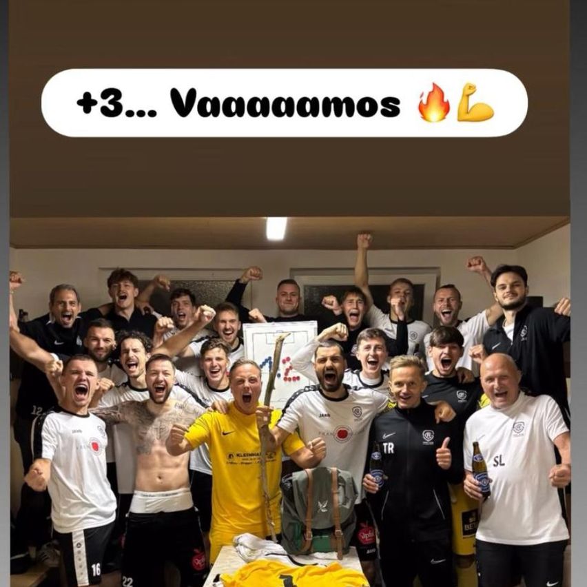 Eine Gruppe von Fußballspielern feiert in einem Raum, wobei ein Spieler einen Pokal hält und ein anderer ein Bier. Im Hintergrund befindet sich ein weißer Banner mit dem Text +3... Vaaaaaamos.
