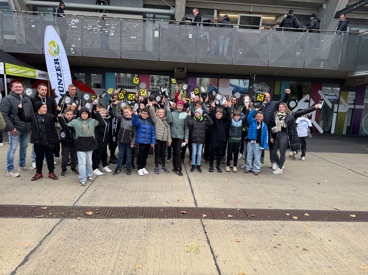 Eine Gruppe von Kindern und Erwachsenen posiert für ein Foto vor einem Gebäude mit Balkon und Geländern. Sie halten gelbe Schilder mit einem 'X' darauf.