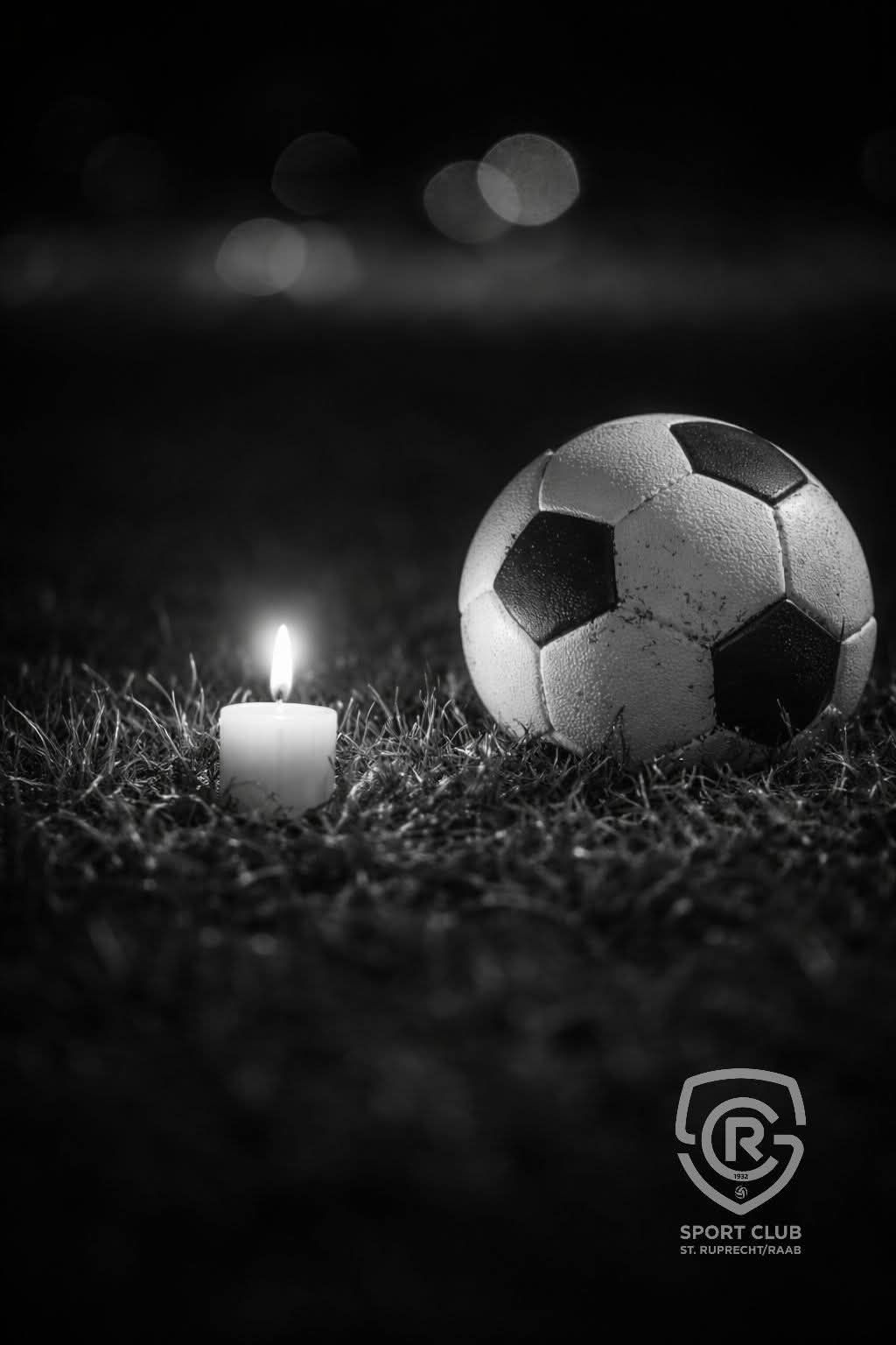 Eine Kerze brennt neben einem Fußball auf einem Rasenfeld in der Nacht. Der Ball befindet sich rechts, während die Kerze links steht.