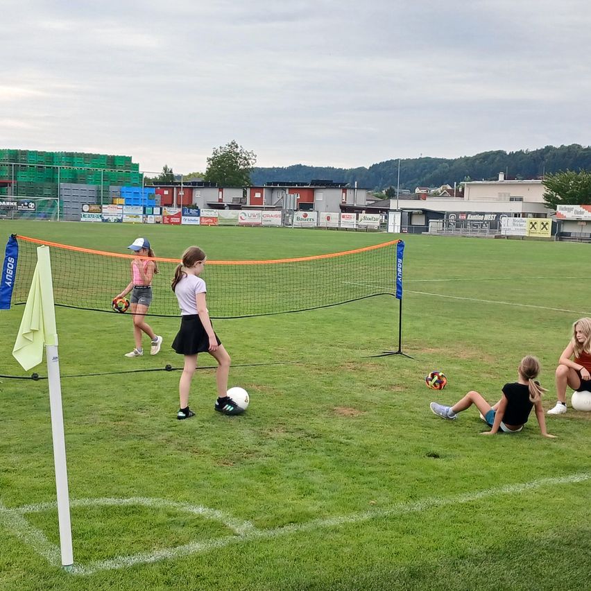 Four girls play soccer on a grassy field with a net in the middle. One girl is holding a soccer ball, and another is sitting on the ground.