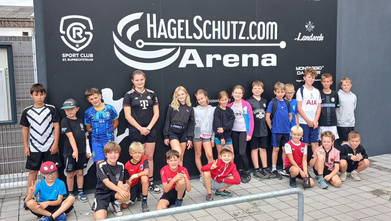 A group of children and young adults are posing for a photo in front of a wall with the logo of Hagelschutz.com Arena. They are wearing different colored jerseys.