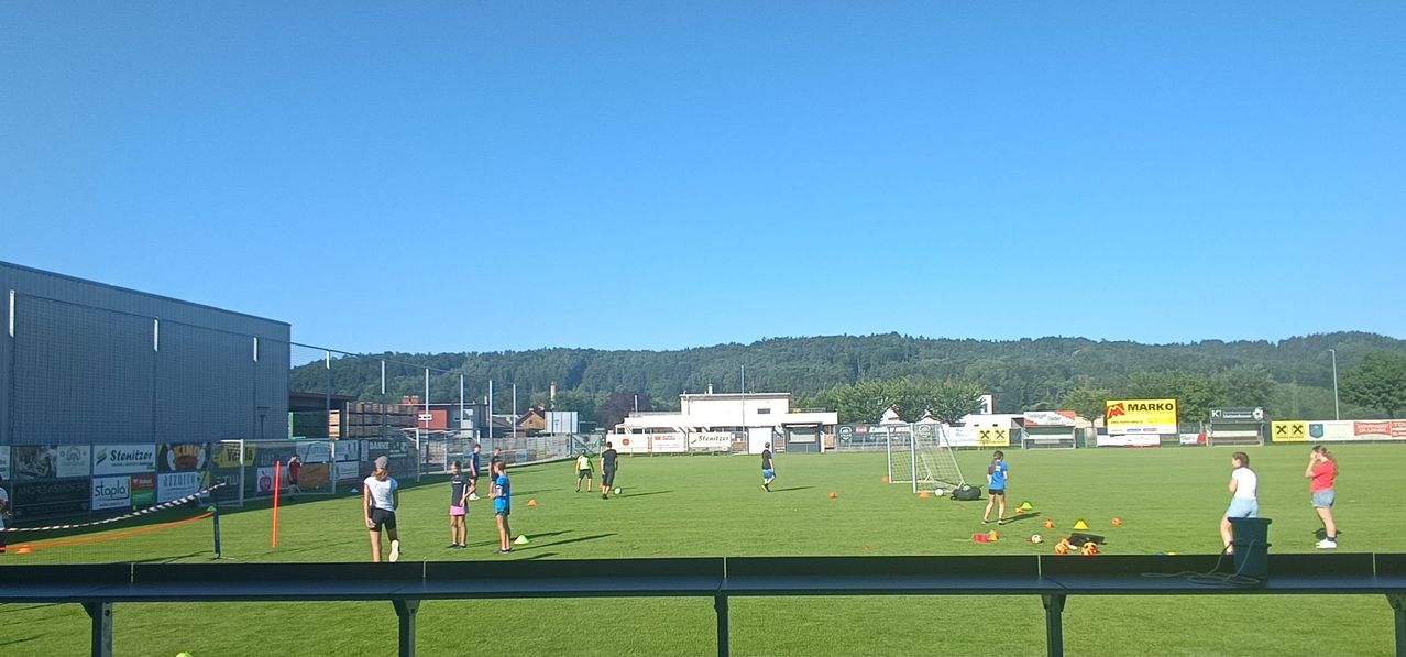 A group of people are practicing soccer on a field with orange cones and a goal. The sky is clear and blue.