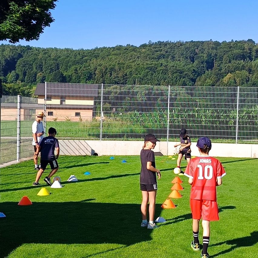 A group of children in sports uniforms are practicing soccer drills on a field with orange cones and a fence in the background.