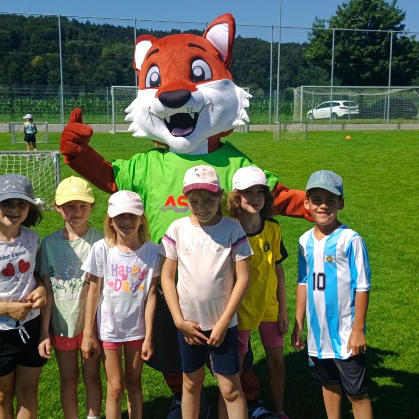 A group of children and a mascot dressed as a fox pose for a photo on a soccer field.