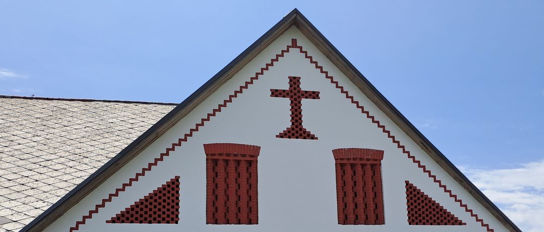 Die Vorderseite eines Kirchengebäudes mit einem dreieckigen Dach. Es zeigt ein rotes Ziegelkreuz-Design über zwei Ziegelfenstern.