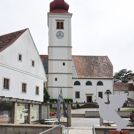 Der Außenbereich einer Kirche mit einem Glockenturm. Vor der Kirche steht eine weiße Taubenstatue.