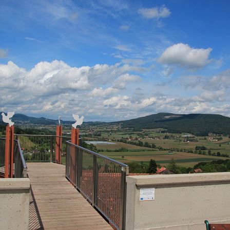 Ein Balkon mit Holzboden und Metallgeländern. Zwei weiße Vögel sitzen auf den Geländern. Der Blick umfasst ein Tal mit Häusern, Bäumen und einem Berg in der Ferne.