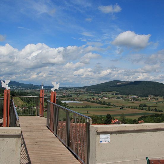 Ein Balkon mit Holzboden und Metallgeländern. Zwei weiße Vögel sitzen auf den Geländern. Der Blick umfasst ein Tal mit Häusern, Bäumen und einem Berg in der Ferne.