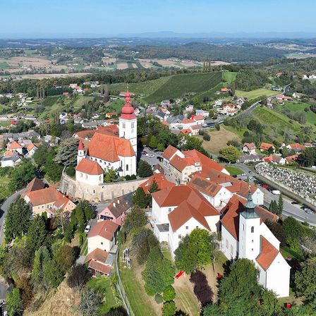 Luftaufnahme einer Stadt mit einer Kirche und mehreren Gebäuden mit roten Dächern, umgeben von Grün und Bergen in der Ferne.