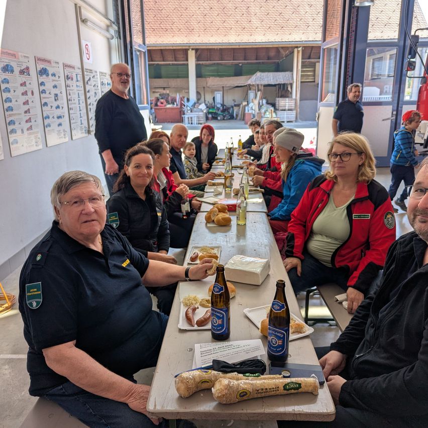 Eine Gruppe von Menschen sitzt an einem Tisch mit Essen und Getränken, möglicherweise in einer Feuerwache. Ein Mann steht hinter dem Tisch, und ein anderer Mann geht vor ihnen. Der Tisch hat Brot, eine Box und Flaschen.