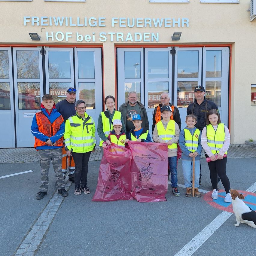 Eine Gruppe von Menschen in gelben Westen und Sicherheitsausrüstung steht vor der Feuerwehr mit einem großen rosa Abfallsack.