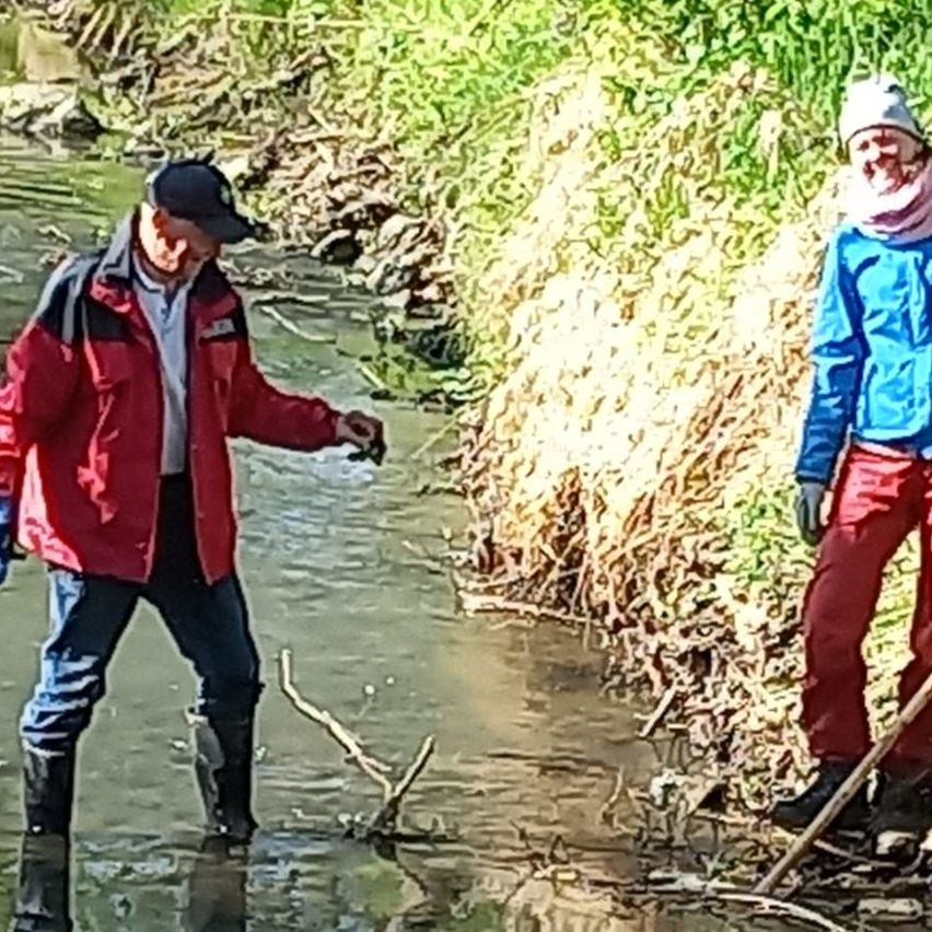 Zwei Personen stehen in einem kleinen Fluss, einer trägt eine rote Jacke und der andere eine blaue. Beide tragen Stiefel und Handschuhe.