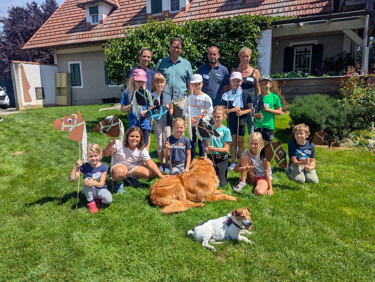 Eine Gruppe von Menschen, darunter Kinder und Erwachsene, posiert für ein Foto auf einem Rasen mit zwei Hunden, einem liegenden und einem stehenden. Die Kinder halten Papierpferde und Fahnen. Ein Haus mit einem Ziegeldach ist im Hintergrund.