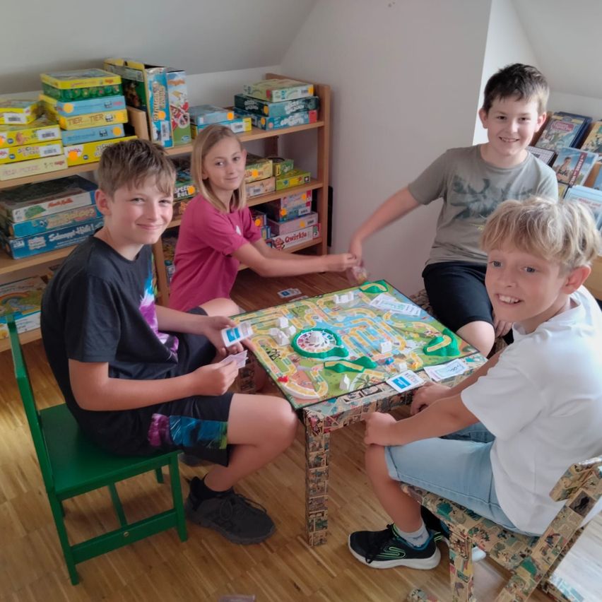 Vier Kinder sitzen an einem Tisch und spielen ein Brettspiel in einem mit Spielzeug gefüllten Raum.