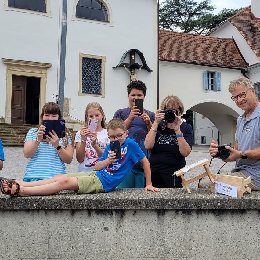 Mehrere Personen sitzen auf einer Betonbarriere und halten ihre Telefone in der Hand, um die Landschaft zu fotografieren. Ein Mann hält eine Kamera. Hinter ihnen steht ein Gebäude mit einem Kreuz oben.