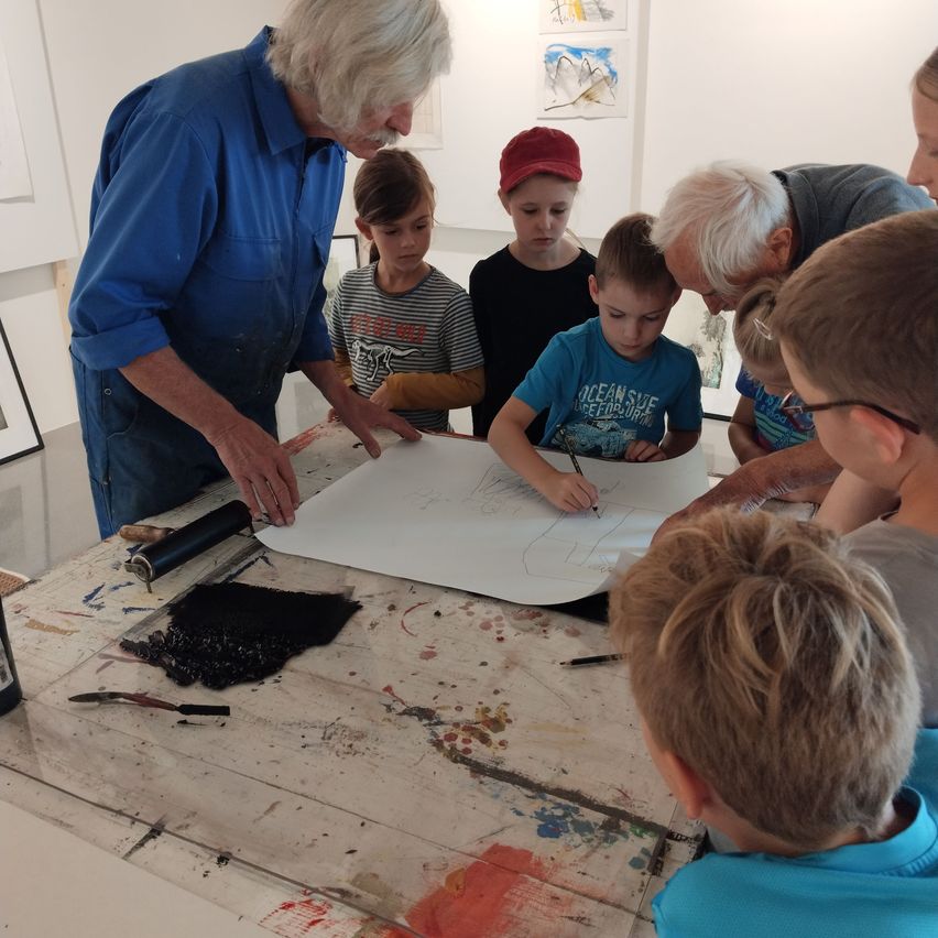 Ein älterer Mann zeigt Kindern, wie man auf einem großen Blatt Papier zeichnet. Sie sind um einen Tisch mit Farbspritzern versammelt. Die Kinder tragen Freizeitkleidung und einige tragen Brillen.