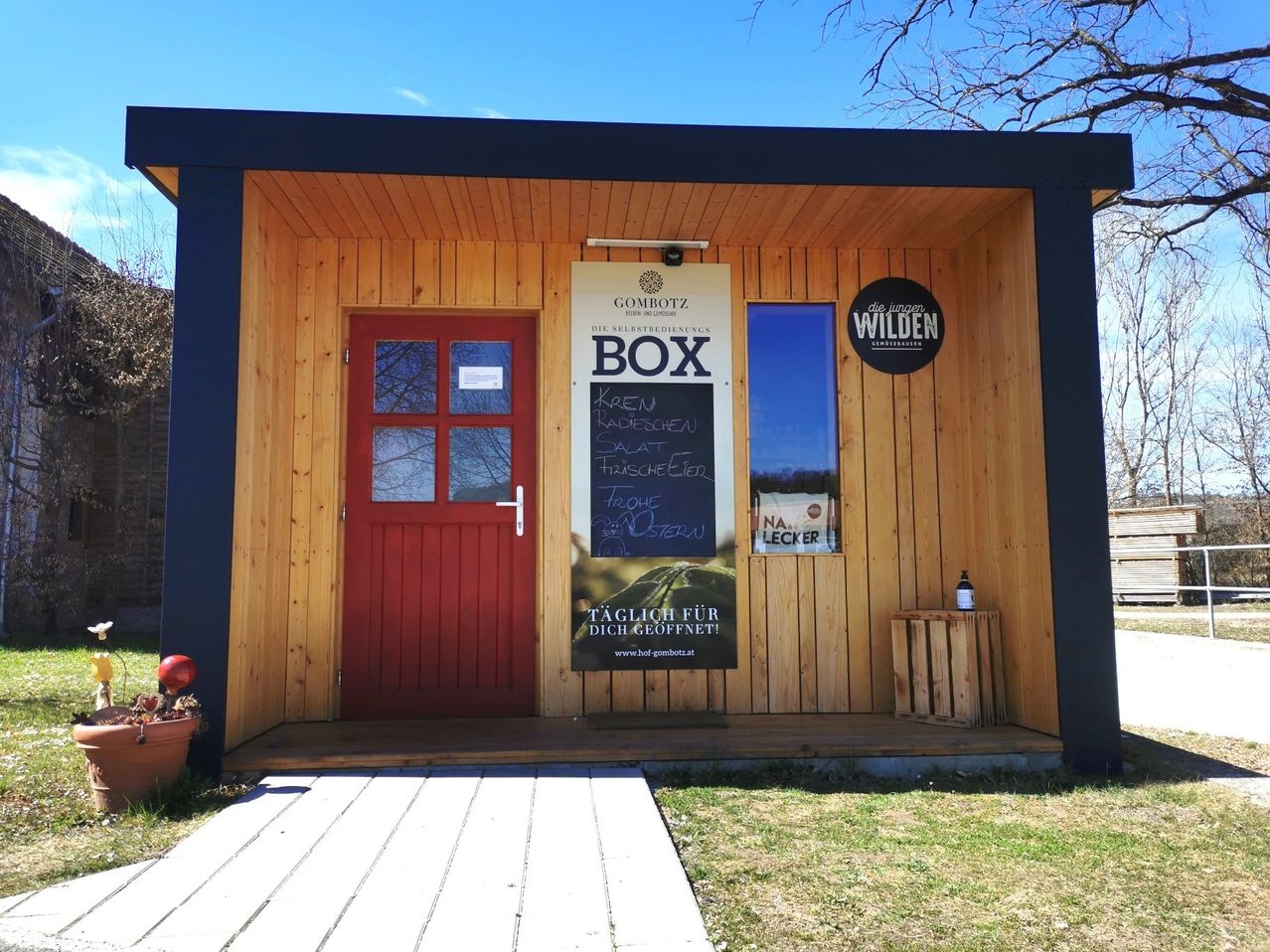 Eine kleine Holzbox mit einer roten Tür steht draußen. Ein Schild an der Wand mit der Aufschrift 'BOX'. Es gibt eine Tafel mit Schriftzug, ein Fenster und eine Flasche auf einem Holzkisten.