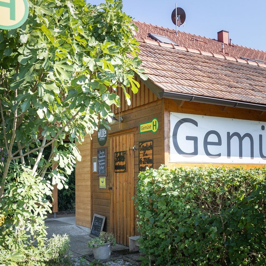 Ein Holzgebäude mit einem Ziegeldach, beschriftet mit 'Gemu', steht vor einem Baum. Das Gebäude hat eine Tür, ein Fenster und ein Schild. In der Nähe befinden sich ein Topf mit Pflanzen und ein Schild mit dem Wort 'Wilde'.