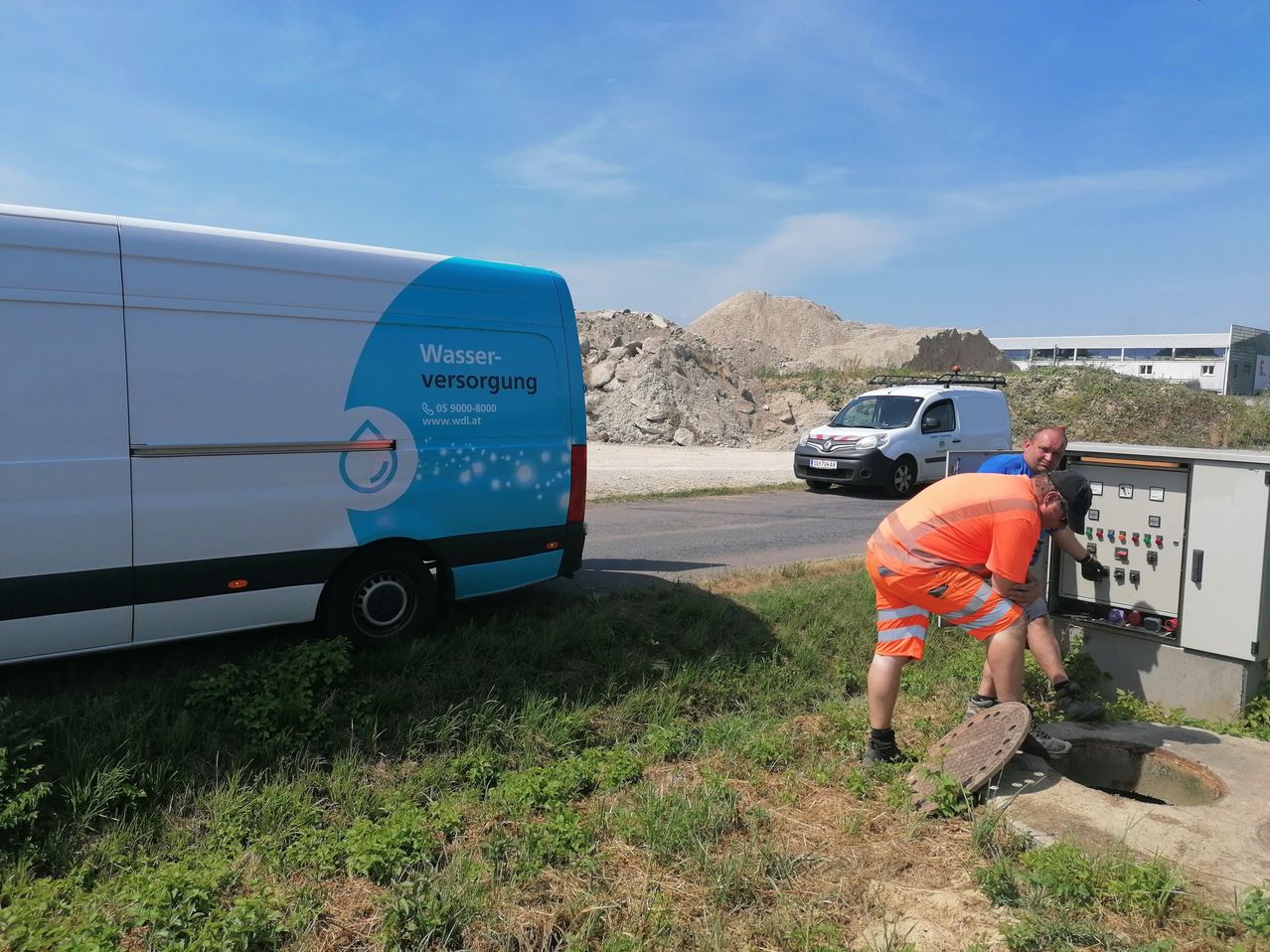 Zwei Arbeiter arbeiten an einem Wasserversorgungssystem in der Nähe eines Lieferwagens und ein weißer Lieferwagen ist am Straßenrand geparkt.