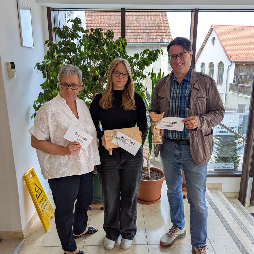 Drei Menschen stehen in einem Gebäude, lächeln und halten Urkunden. Eine Frau links und ein Mann rechts tragen eine Brille. Die Frau in der Mitte hält eine kleine Holzkiste.