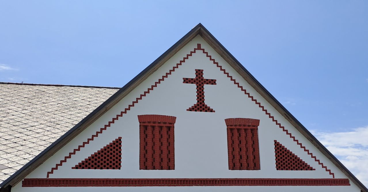 Ein weißes dreieckiges Gebäude mit einem roten Kreuz und zwei roten Ziegelfenstern. Das Dach ist braun.