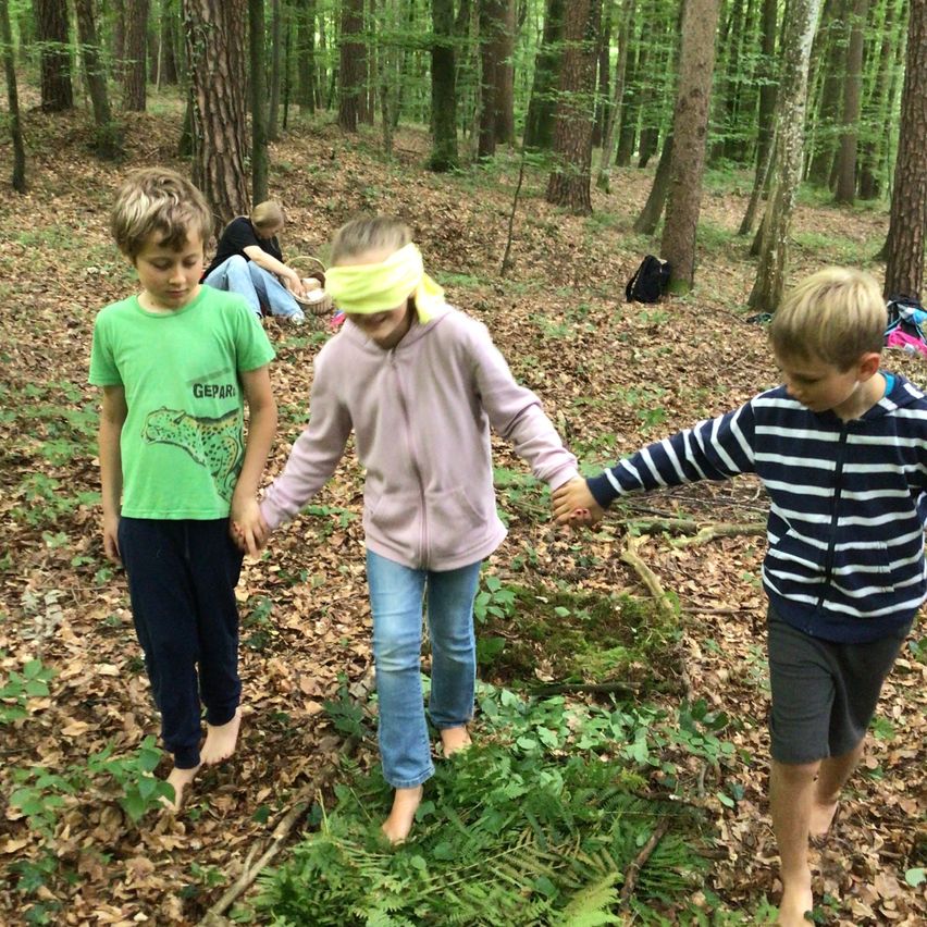 Drei Kinder gehen barfuß durch einen Wald, eines mit verbundenen Augen, während eine Frau im Hintergrund beobachtet.