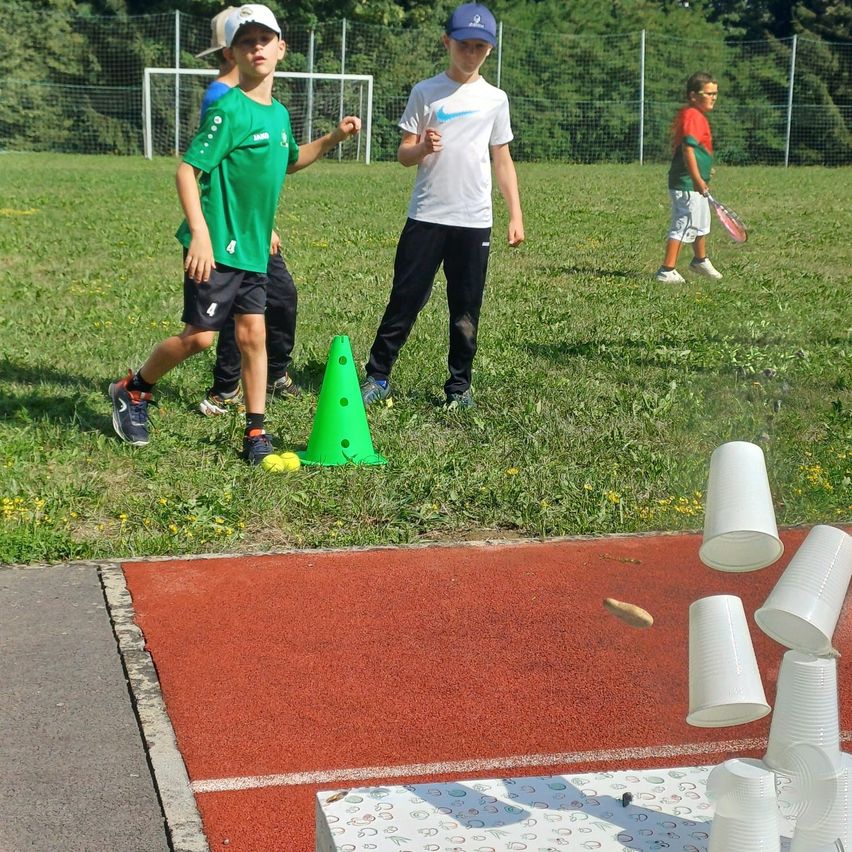 Kinder spielen auf einem Grasfeld mit Bechern und einem Kegel. Ein Junge trägt ein grünes Shirt. Ein anderer Junge trägt ein weißes Shirt und einen blauen Hut.