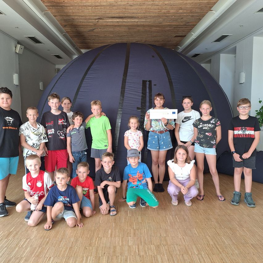 Eine Gruppe von Kindern und Erwachsenen posiert für ein Foto vor einem großen Zelt, einer hält ein Papier mit einem Logo.