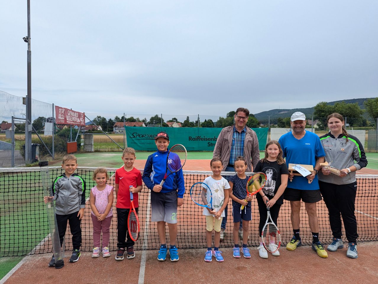 Eine Gruppe von Kindern und Erwachsenen steht auf einem Tennisplatz, hält Tennisschläger und lächelt für ein Foto. Dahinter befindet sich ein Netz, ein Banner und ein klarer Himmel.