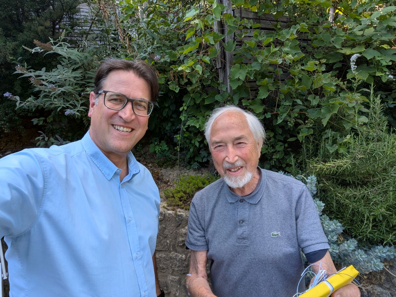 Zwei Männer, einer mit Brille und hellblauem Hemd, der andere mit weißem Bart und grauem Poloshirt, stehen in einem Garten.