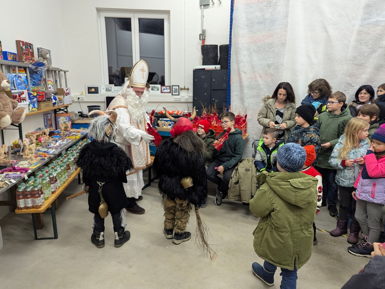 Eine verkleidete Figur interagiert mit Kindern in einem Raum. Dahinter steht ein Tisch mit Spielzeug, und Erwachsene beobachten. Der Raum hat ein Fenster und Aufbewahrungsboxen.