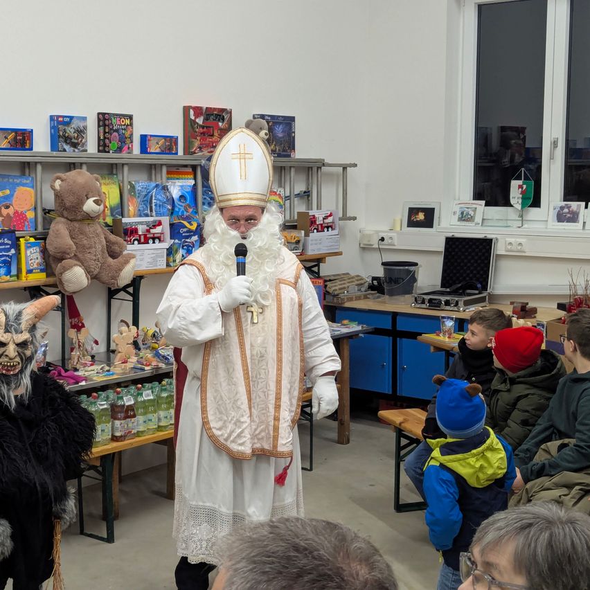 Ein Mann, der als Papst verkleidet ist, spricht in ein Mikrofon in einem Raum voller Kinder. Ein Regal mit Spielzeug und Büchern steht hinter ihm, und ein Tisch mit Flaschen und einem Eimer befindet sich in der Nähe.
