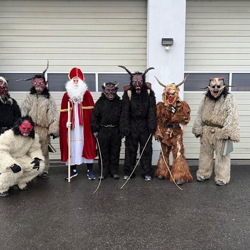Eine Gruppe von Menschen in festlichen Kostümen posiert vor einem Gebäude, darunter der Weihnachtsmann und andere in Dämonenmasken.