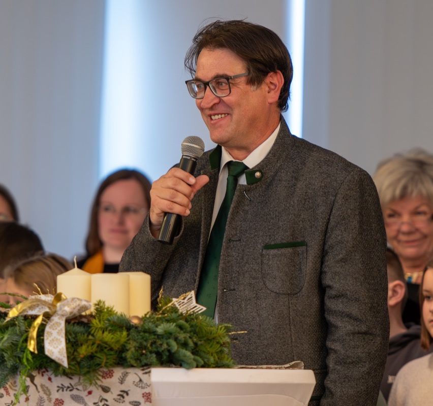 Ein Mann mit Brille steht und lächelt, hält ein Mikrofon in einer formellen Umgebung mit einem Kranz und Kerzen. Hinter ihm beobachtet eine Menge von Menschen.