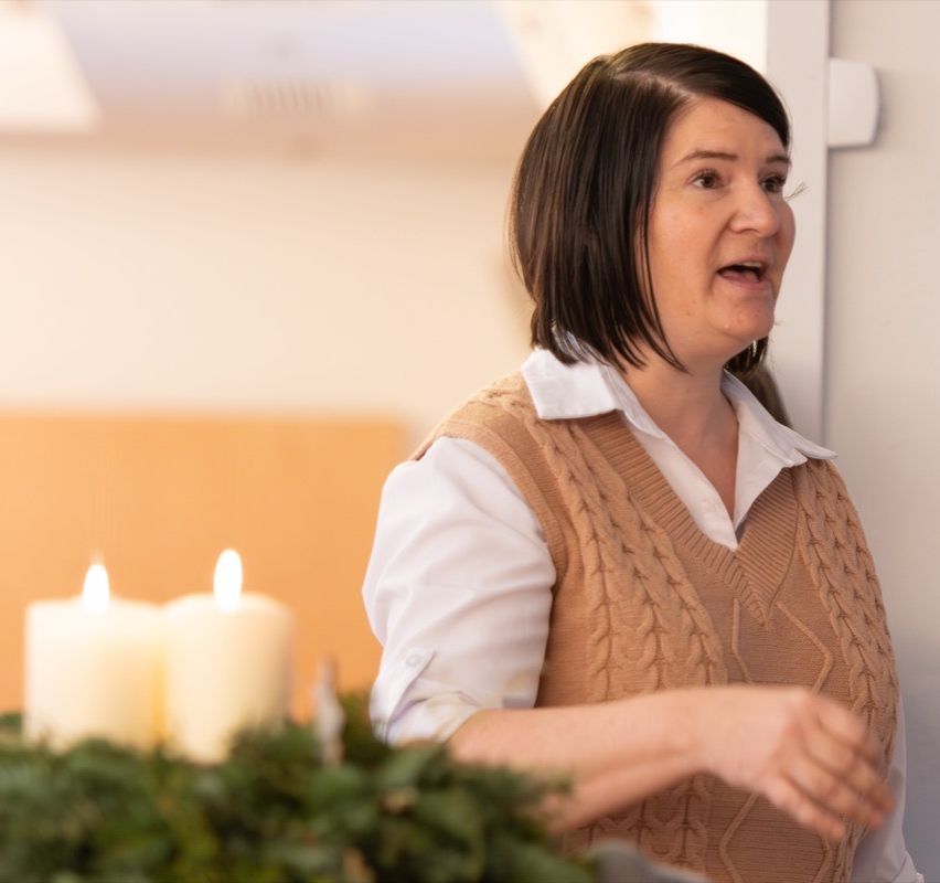 Eine Frau mit kurzen Haaren und einem gestrickten Westchen steht vor brennenden Kerzen und Grünpflanzen und spricht.