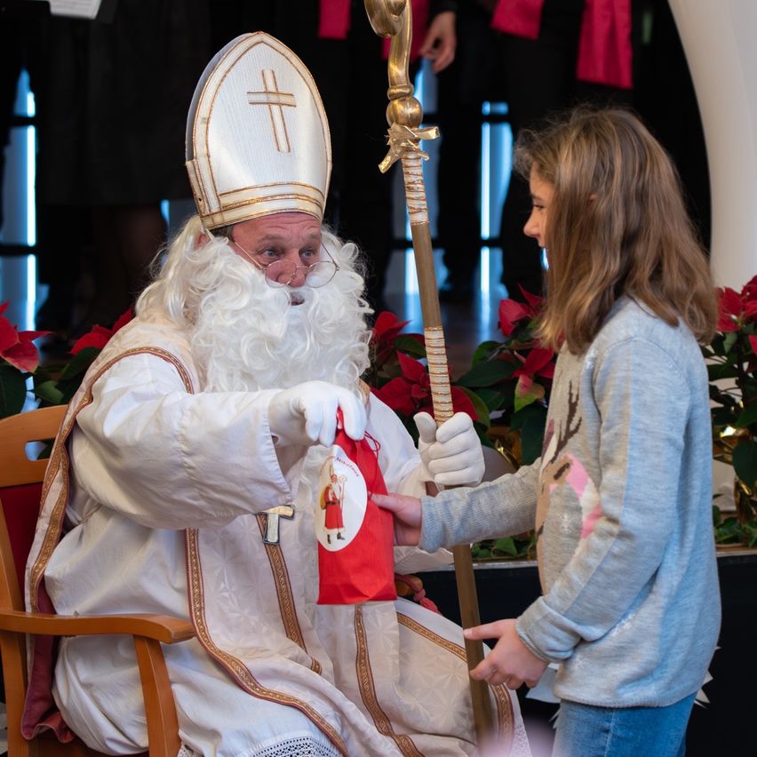 Eine als St. Nikolaus verkleidete Person gibt einer jungen Mädchen in einer Kirchenumgebung einen roten Beutel.