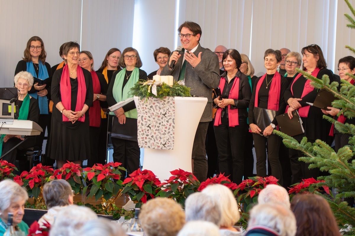 Ein Mann hält eine Rede an einem Podium vor einer Menge, mit Frauen in Schals hinter ihm. Das Podium hat eine Blumenarrangement und Kerzen.