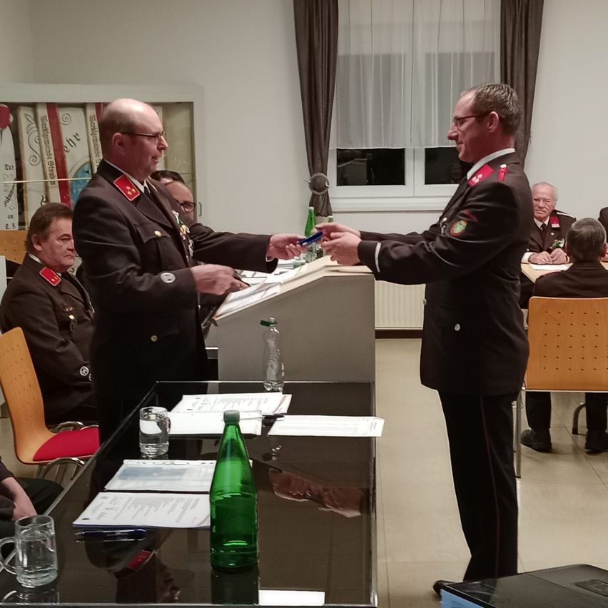 Zwei Männer in Militäruniformen tauschen Dokumente an einem Podium in einem Konferenzraum aus. Hinter ihnen sitzen weitere Personen auf Stühlen. Papiere und Flaschen sind auf dem Tisch.